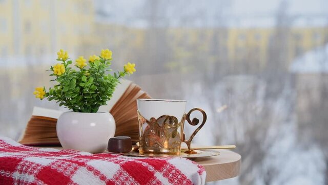 Golden coffee cup, book and vase with flowers on table in cafe. Winter time, It snows outside. Coziness atmosphere