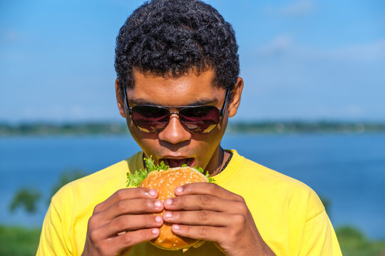 Hungry African American man enjoying the taste of hamburger outdoor in summer. Concept of love to junk food diet 