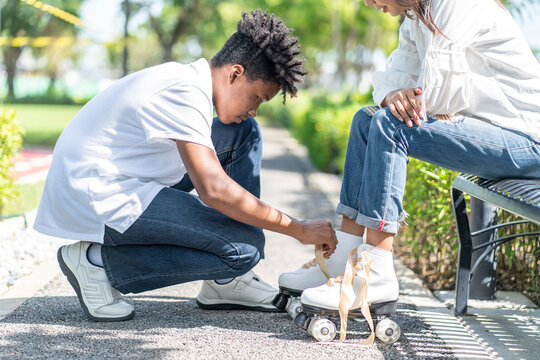 An African-American teenager helps an Asian woman to tie a roller skate to an Asian woman to practice roller skating in the park. 