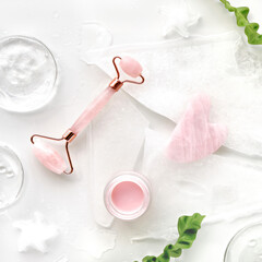 Pink quarts face roller, gua sha stone, pink cream in glass jar. Off white flat lay in green and pink with ice and exotic fern leaves. Beauty facial massage. Natural cosmetics laboratory.