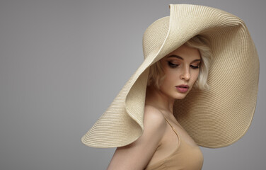 Blonde girl in a large wicker beach hat with wide brim. Grey background.