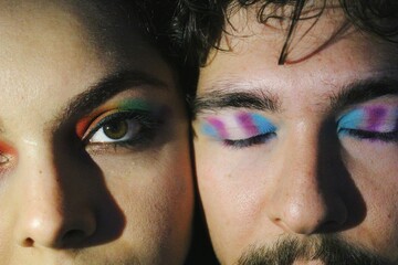 portrait of closeup faces of two lgbtq queer friends faces with rainbow pride flag and trans pride flag makeup / one eye open version - obrazy, fototapety, plakaty