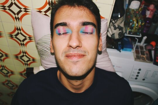 portrait face of lgbt queer young man smiling with pink blue & white colored trans pride flag makeup with flash at night