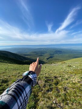 Hand of girl in plaid shirt standing on top of mountain points to clear blue sky with clouds. Horizontal image. Concept of travel. Beautiful mountain landscape. Mobile photography.