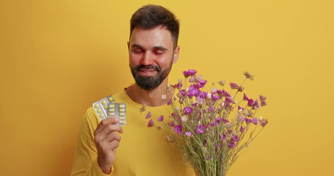 Allergy treatment concept. Happy man takes pills against hypersensitivity can smell wildflowers enjoys pleasant scent has red swollen eyes as reaction to allergen isolated over yellow background