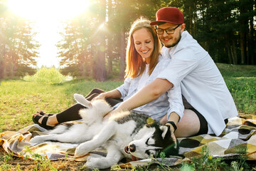 A young couple, a man and a woman, are stroking the stomach of their Husky dog while sitting on a blanket. Walking a pet in the park to a clearing. The concept of happiness, love, joy.