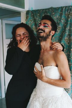 portrait of queer young man with beard & body hair in white bridal gown & young woman in black suit laughing in home interior