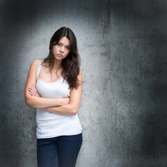 Outdoor portrait of a sad looking young woman wearing a white top and blue jeans in front of a dark wall, copy space on the right side of the image