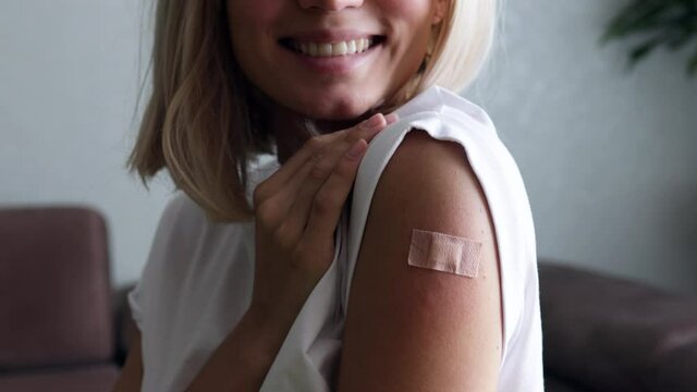 Portrait of a female smiling after getting a vaccine. Woman holding down her white shirt sleeve and showing her arm with bandage after receiving vaccination. Concept of recommended inoculation