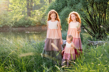 three red-haired sisters in long linen dresses are resting on lake in the park on a sunny summer day.