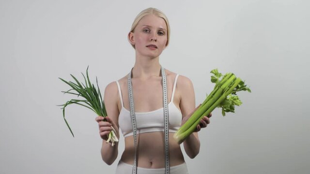 A slender woman with a measuring tape around her neck holds green onions and celery in her hands. Healthy lifestyle, diet promotion. Isolated on white background, slow motion, HD.