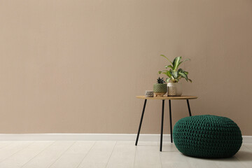 Knitted pouf and table with houseplants near beige wall indoors. Space for text