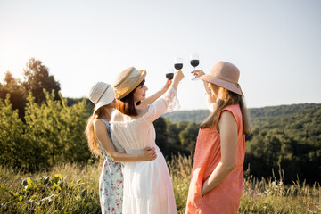 Back view of three stylish stunning pretty girlfriends standing on green grass and raised the glasses of red wine, celebrating party. Women on the nature picnic.