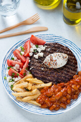 Blue and white plate with serbian traditional pljeskavica, red tomatoes, fries and prebranac, vertical shot on a white stone background