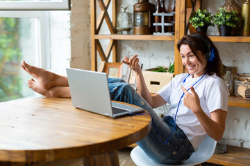 Happy smiling woman in blue headphones in front of a laptop monitor at home. She puts her feet on...