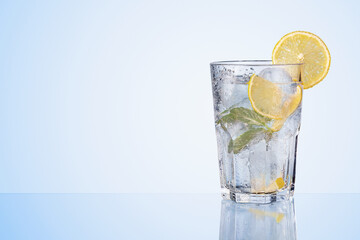 Glass of water with fresh lemons, leaves of mint and ice cubes on blue background. Refreshing lemonade, lemon drink.
