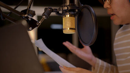 Asian female podcaster recording and broadcasting her podcast from home. Woman working from home at night reading a notepad, paper work and talking into a microphone.