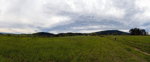 Rabka Zdroj, Poland : Panorama view of a person with backpack hiking or walking in a middle of a meadow field in country side against hills near rabka on a way to lubon wielki - obrazy, fototapety, plakaty