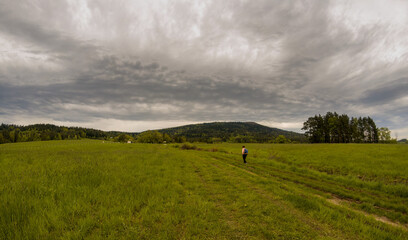 Rabka Zdroj, Poland: Panorama view of a person hiking or walking in a middle of a meadow field in country side against hills near rabka on a way to lubon wielki - obrazy, fototapety, plakaty