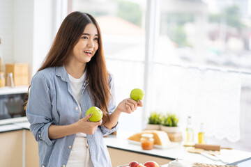 Young Asian women with casual dress in the morning and eat fresh apple fruit for breakfast in the kitchen with different type of vegetable and healthy food. Good nutrition eating human body condition.