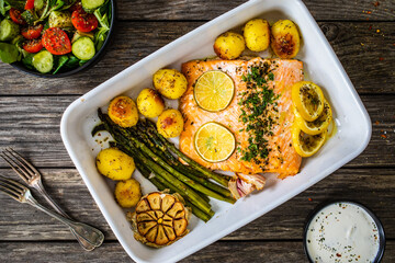 One pot dinner - roasted salmon steak with asparagus, lemon, potatoes, parsley and garlic in cooking pot on wooden table
