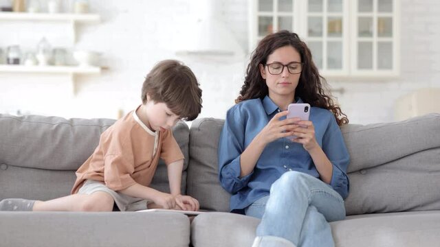 Smartphone addicted mother use cellular gadget sit together with small son playing games on digital tablet on sofa. Young mom text or surfing internet in mobile app and kid with touchscreen pc at home