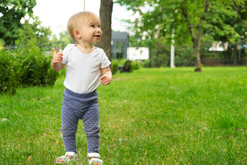 Adorable happy blonde caucasian baby girl of 1 year old, standing on lawn, laughing, smiling.Infant, spending spare time in summer outdoors in park. Child and nature concept.Copy space for text