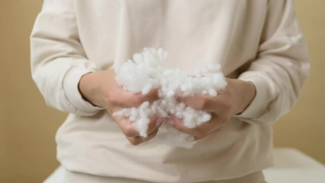 Medium shot. A girl in a vanilla-colored hoodie on a yellow background tears up a voluminous artificial filler in the form of white balls