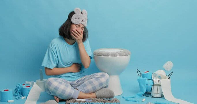 Asian woman suffers from abdominal pain feels unwell after hangover has nausea and vomits in toilet keeps hands on stomach wears pajama and sleepmask cannot go to sleep spends night in restroom.
