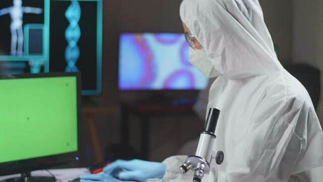Asian Doctor In Protective Suit Working With Mock Up Green Screen Computer Display In A Modern Lab. Laboratory Assistant Workplace
