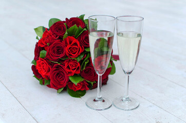 Two wedding glasses with champagne and bridal bouquet with red roses on a white wooden background. Close up. Wedding ceremony, celebration. Wedding attributes, symbol. Bouquet for Valentine's day