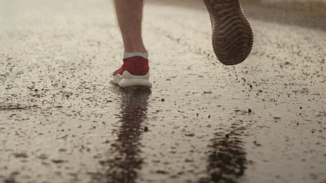 Men's feet in sneakers stand on the wet asphalt in rain and start running through puddles, closeup, back view