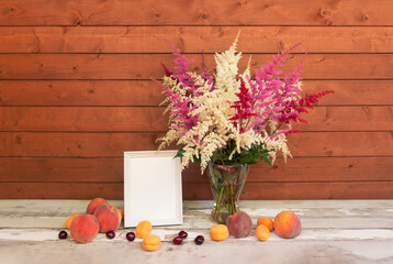 White, red and pink beautiful astilbe flowers in glass vase, white frame and fresh fruits on aged wooden table.