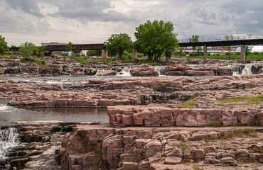 Sioux Falls, SD, USA - June 2, 2008: Foot and bike bridge over the cascade of waterfalls with brown-beige rock field under heavy gray sky. Some green foliage.