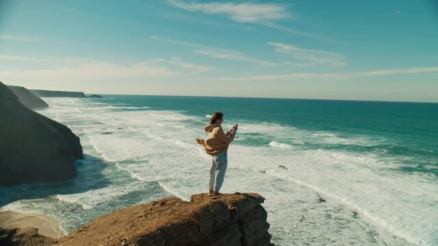 Woman in free flowing outfit stand on edge of epic picturesque cliff, record landscape views on video on smartphone. Cinematic adventure destination and travel blogger concept