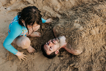 Young girl buries dad in the sand on Waikiki beach during sunset