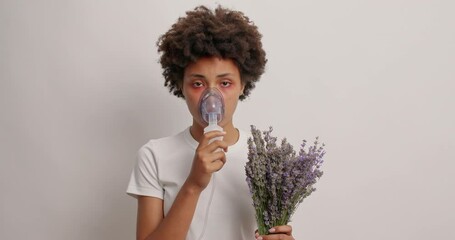 Allergic curly woman has asthmatic attack being allergic to lavender breathes with help of nebulizer chamber has red watery eyes isolated over white background. Symptoms of seasonal disease.