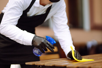 Disinfecting to prevent COVID-19. Waiter in protective face mask and gloves cleaning and disinfecting restaurant table for next customer.