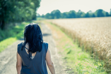 Young woman with black hair is walking along a dirt road between forest and wheat field in sunny weather