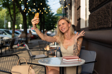 A blonde with a tattoo on her arm sits in a summer cafe and emotionally talks on the phone via video communication. Blogger concept