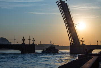 Fototapeta na wymiar a dark silhouette of the open Trinity Bridge and a warship passing through it against the background of a beautiful golden dawn and the rising sun in Saint-Petersburg Russia