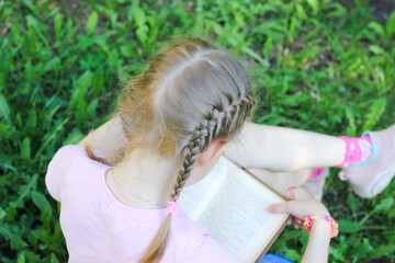 little blonde girl with pigtails in the summer outdoors in the greenery reads a book, photo from the back