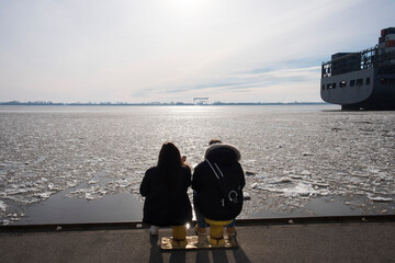two silhouettes of unrecognizable people on the banks of the frozen Elbe in winter in Hamburg and a container ship