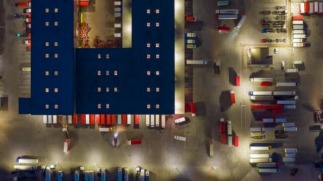 Aerial top down view hyper lapse (hyperlapse - motion time lapse) of a logistics park with a loading hub. Semi-trailer trucks stand at warehouse ramps for load and unload goods at night. Zoom out 