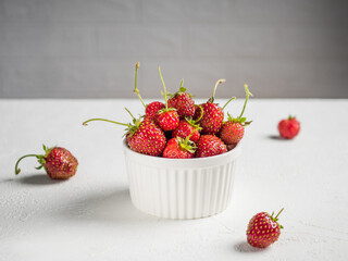 Appetizing fresh strawberries in a white bowl on a white concrete background. The red berries lay beautifully around the file. White decorated brick in the background. Vegan diet. Horizontally
