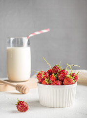 Appetizing fresh strawberry with green cuttings lies in a white bowl on a white background. A glass of milk with a straw in the background. Breakfast concept. Home comfort. Vertical