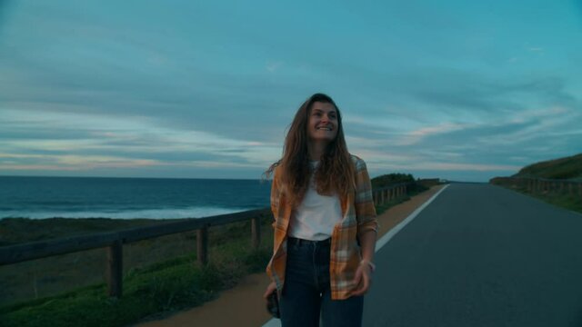 Happy and excited young woman photographer walk on cinematic empty road on ocean coastline. Make photos of sunset and landscape. Millennial woman make create content for social media or memories