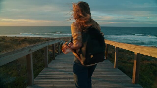 Camera follows happy young woman run on wooden boardwalk to ocean at sunset. Inspirational wanderlust road trip adventure. Authentic and real emotions of discovery and travel. Millennial traveller 