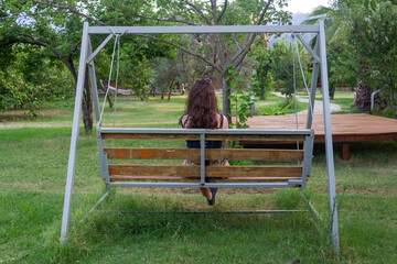 Backward view of a woman with long hair sitting alone on a wooden swing bench in the garden full of green trees. Selective focus. 