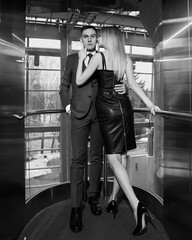 Fototapeta na wymiar Young stylish fashionable caucasian couple hugging and kissing in hotels elevator. Handsome man in classical suit and blonde pretty woman in black dress